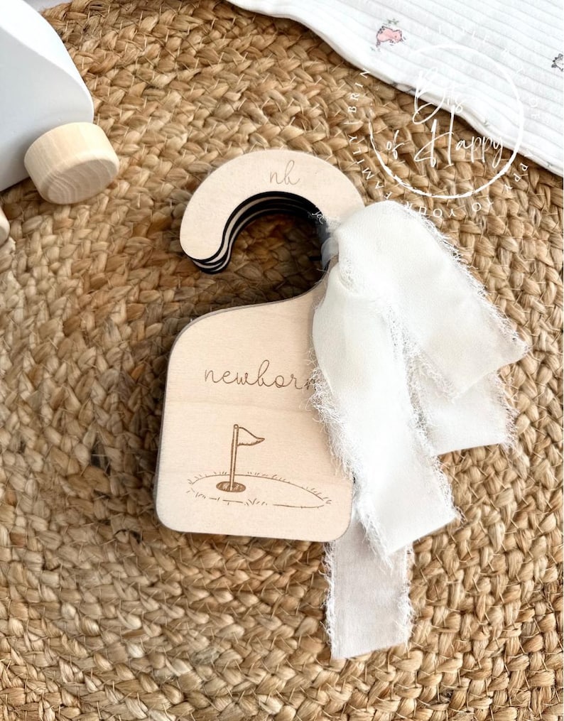 May include: A wooden baby clothes hanger with the word "newborn" and a golf flag design. The hanger has the initials "nh" on the top and is adorned with a white ribbon. The background is a woven, natural-colored material.