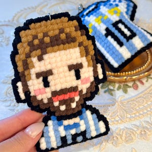 May include: A pixel art style yarn keychain featuring a person with brown hair and a beard, wearing a light blue and white striped jersey. A second matching jersey keychain with the number 10 is visible in the background, both crafted with yarn.