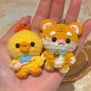 Può includere: Due portachiavi all'uncinetto fatti a mano: un pulcino giallo con cresta arancione e fiocco blu, e una tigre arancione e bianca con collare blu e campanella dorata. Entrambi hanno occhi neri e sono tenuti in mano.