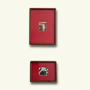 May include: Two rectangular wooden frames with a dark brown finish, each featuring a vibrant red mat board. The top frame contains a small vertical photograph, while the bottom frame holds a small horizontal black and white photograph.