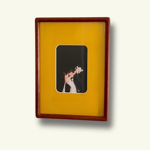 Puede incluir: Un marco de fotos rectangular con acabado en madera oscura y un paspartú de color amarillo vibrante. El marco contiene una fotografía vertical centrada de dos personas con ropa formal abrazándose sobre un fondo negro.