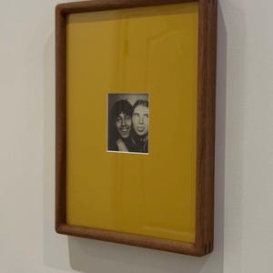 May include: A framed photograph featuring two people in a black and white image. The frame is made of wood and has a rectangular shape. The background of the frame is yellow. The photograph is mounted on a white wall.