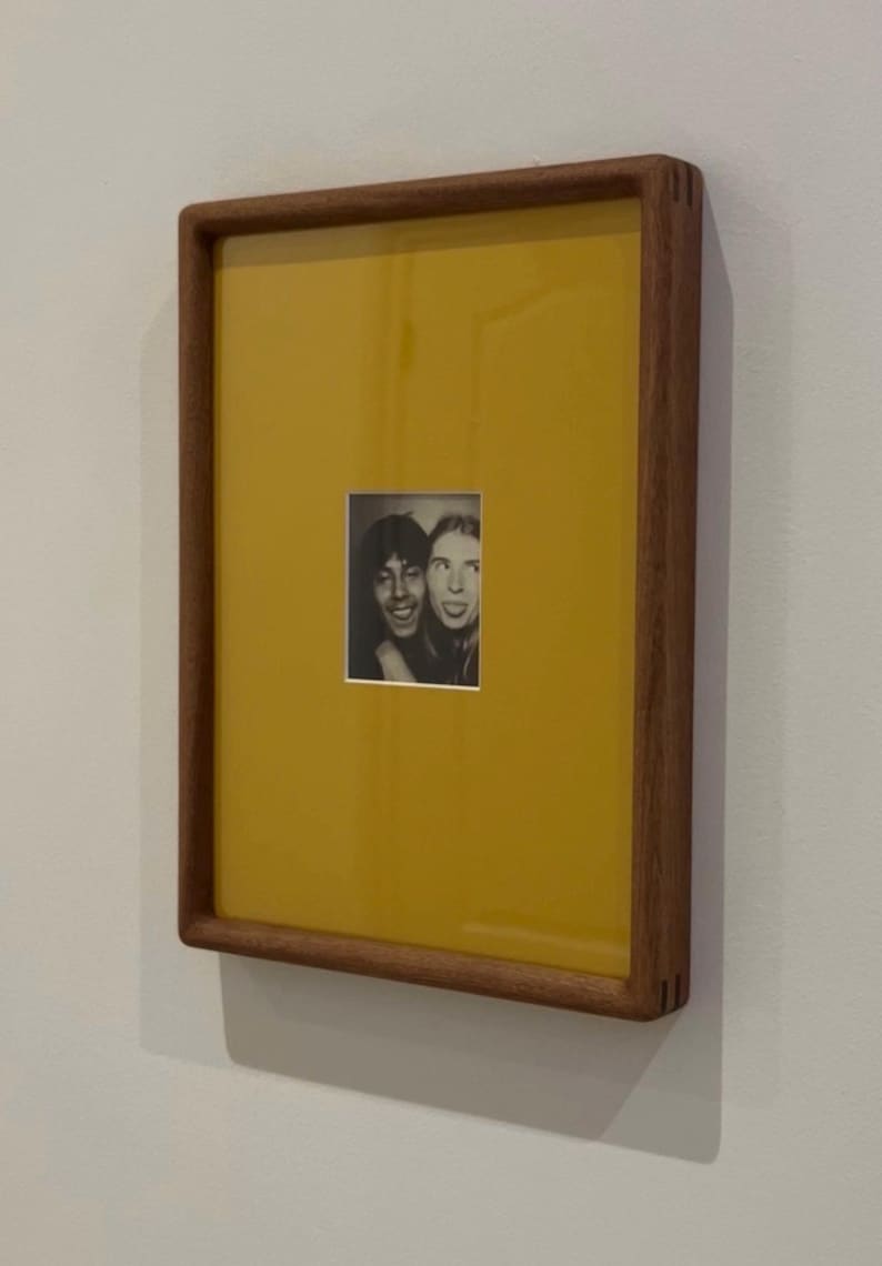 May include: A framed photograph featuring two people in a black and white image. The frame is made of wood and has a rectangular shape. The background of the frame is yellow. The photograph is mounted on a white wall.