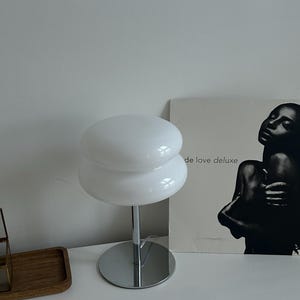 May include: A white, mushroom-shaped table lamp with a chrome base and stem. The lamp has two stacked, rounded shades. The lamp is on a white surface. A black and white album cover is in the background.