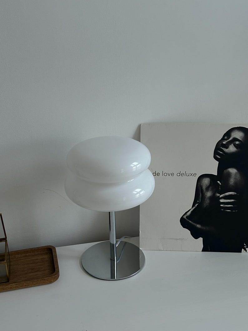 May include: A white, mushroom-shaped table lamp with a chrome base and stem. The lamp has two stacked, rounded shades. The lamp is on a white surface. A black and white album cover is in the background.