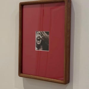 May include: A framed artwork featuring a black and white photograph of a person with their hand over their face, set against a red background. The frame is made of wood and is rectangular.
