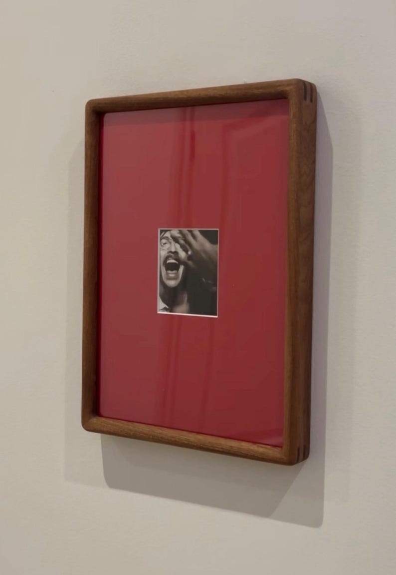 May include: A framed artwork featuring a black and white photograph of a person with their hand over their face, set against a red background. The frame is made of wood and is rectangular.