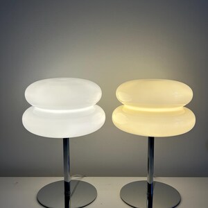 May include: Two modern table lamps with a stacked, rounded design. One lamp is illuminated with a warm glow, while the other is off. Both lamps have a chrome base and stem, and a white, opaque shade.