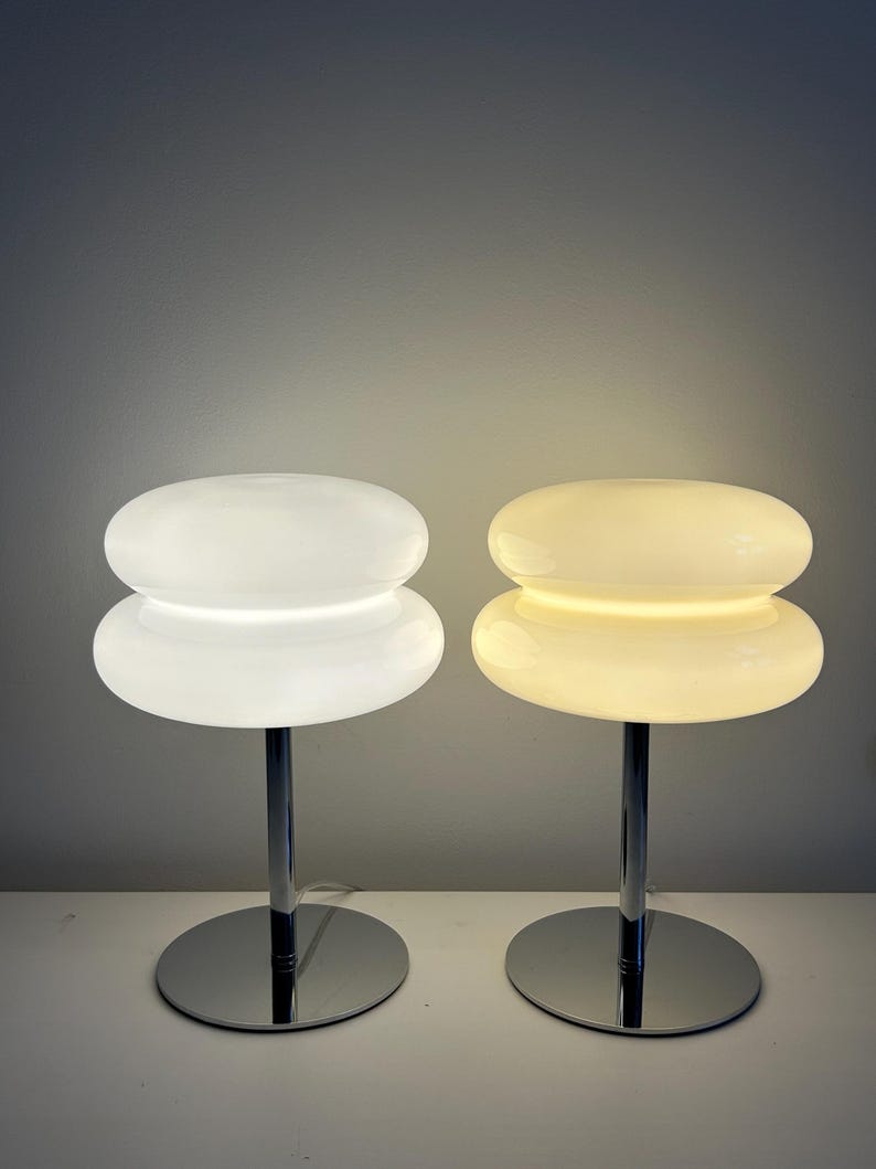 May include: Two modern table lamps with a stacked, rounded design. One lamp is illuminated with a warm glow, while the other is off. Both lamps have a chrome base and stem, and a white, opaque shade.