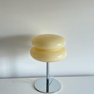 May include: A retro-style table lamp with a stacked, rounded shade in a creamy yellow hue. The lamp features a chrome-finished stem and a circular base. The design is reminiscent of mid-century modern aesthetics, suitable for home decor.