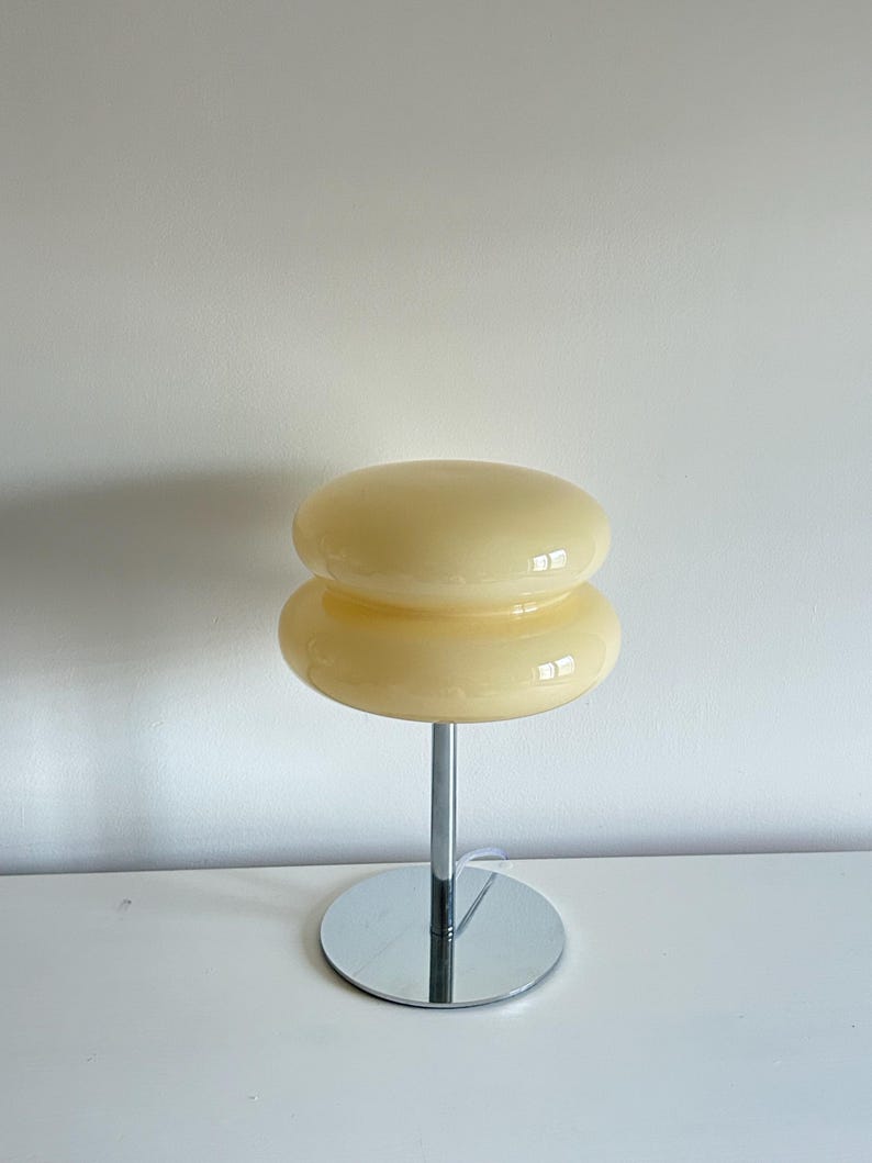 May include: A retro-style table lamp with a stacked, rounded shade in a creamy yellow hue. The lamp features a chrome-finished stem and a circular base. The design is reminiscent of mid-century modern aesthetics, suitable for home decor.