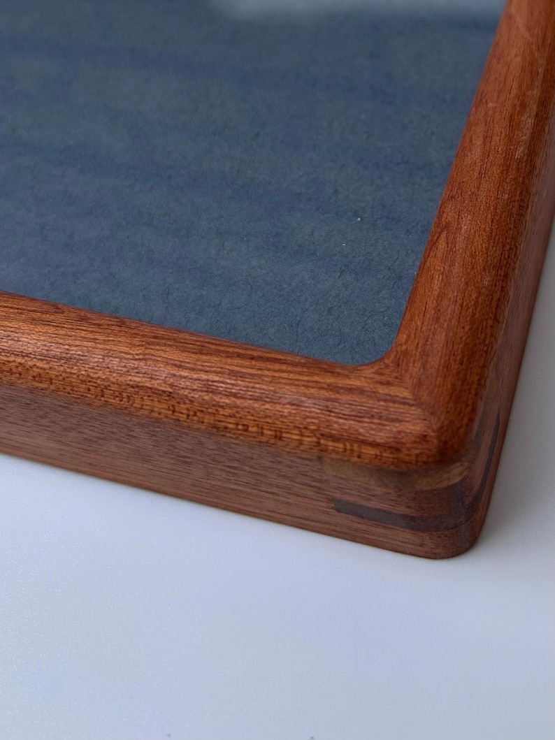 May include: Close-up of a wooden picture frame with a dark blue interior. The frame is made of reddish-brown wood with rounded corners and visible joinery. The frame is on a white surface.