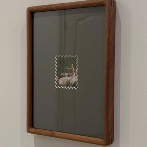 May include: A rectangular wooden picture frame with a dark gray background. A small, postage stamp-like image with a serrated edge is centered on the frame, depicting a scene of people outdoors. The frame is mounted on a white wall.