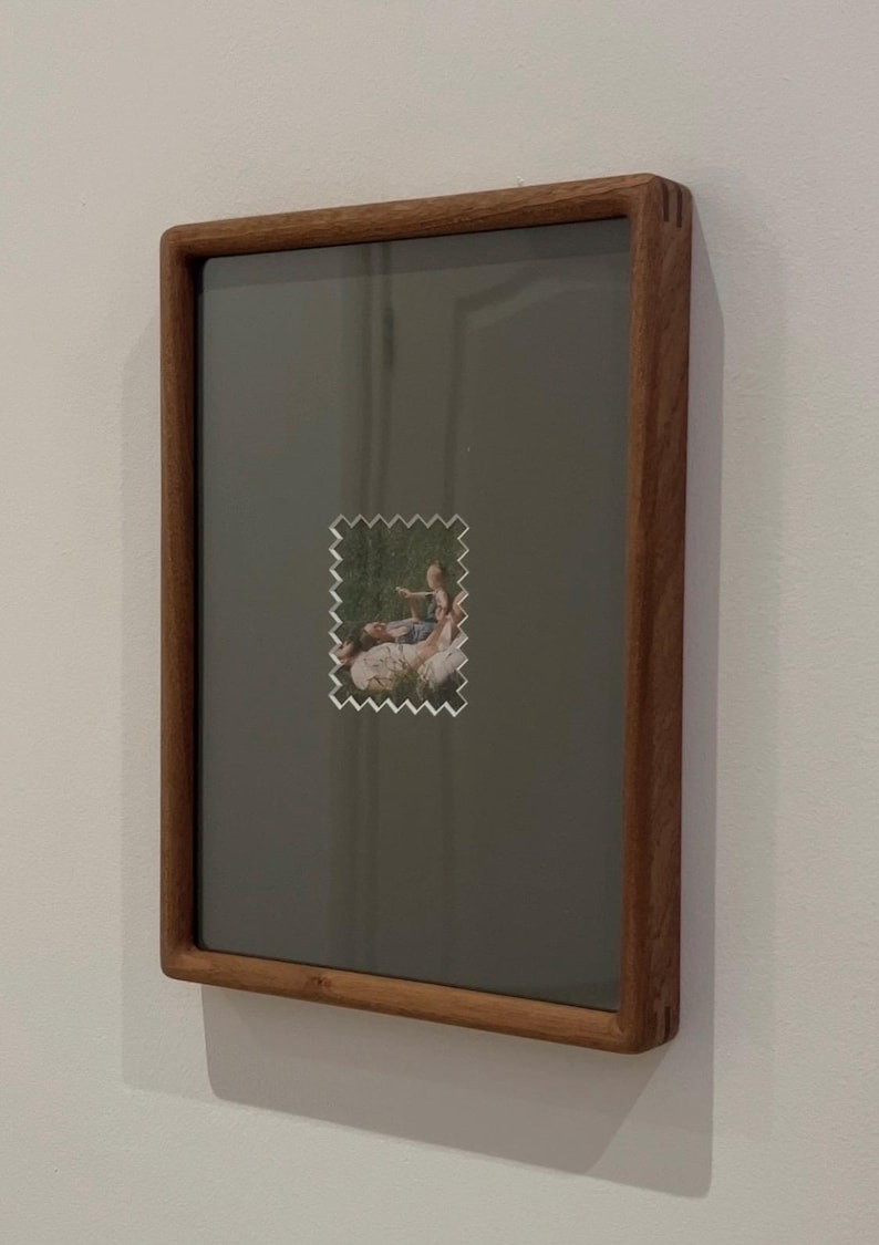 May include: A rectangular wooden picture frame with a dark gray background. A small, postage stamp-like image with a serrated edge is centered on the frame, depicting a scene of people outdoors. The frame is mounted on a white wall.