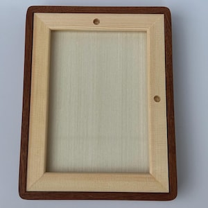 May include: A rectangular wooden frame with a light-colored interior and a dark brown border. The frame has rounded corners and two small holes. The wood grain is visible on the interior and the border.