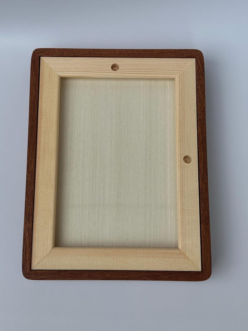 May include: A rectangular wooden frame with a light-colored interior and a dark brown border. The frame has rounded corners and two small holes. The wood grain is visible on the interior and the border.