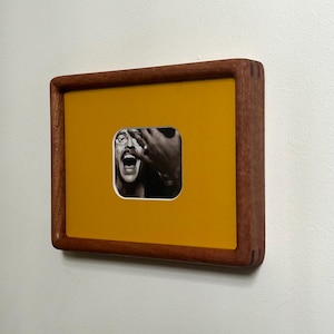May include: A framed artwork featuring a black and white photograph set against a mustard yellow background. The frame is made of dark wood. The photograph depicts a person with their mouth open and hands near their face.