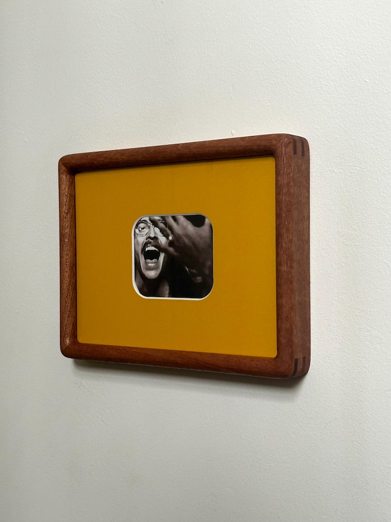 May include: A framed artwork featuring a black and white photograph set against a mustard yellow background. The frame is made of dark wood. The photograph depicts a person with their mouth open and hands near their face.