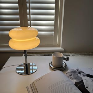 May include: A modern table lamp with a stacked, rounded shade in a warm, off-white color. The lamp has a chrome base and stem. A white mug with a silver handle sits on a silver coaster, next to an open book.