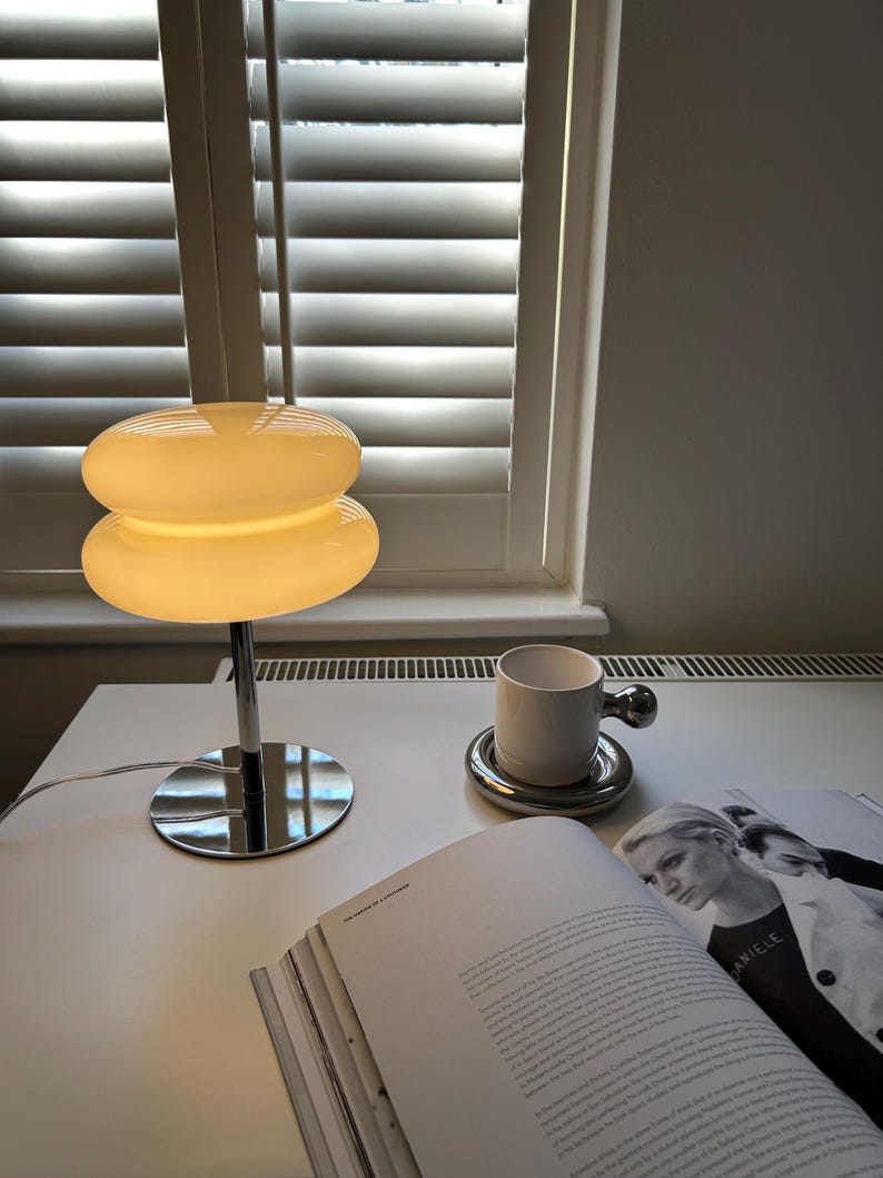 May include: A modern table lamp with a stacked, rounded shade in a warm, off-white color. The lamp has a chrome base and stem. A white mug with a silver handle sits on a silver coaster, next to an open book.