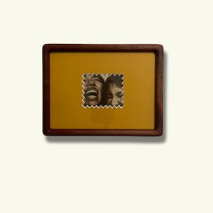 May include: A rectangular wooden picture frame with a dark brown finish. The frame holds a mustard-colored background with a small, postage stamp-like image of two people laughing. The image is in black and white.