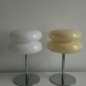 May include: Two modern table lamps. One is white, the other is a pale yellow. Each lamp has a stacked, rounded shade atop a chrome stem and circular base. The lamps are designed for a minimalist aesthetic.