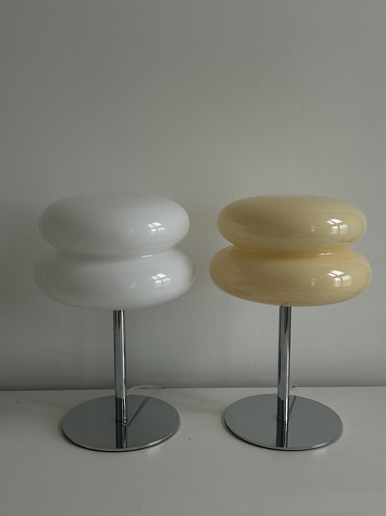 May include: Two modern table lamps. One is white, the other is a pale yellow. Each lamp has a stacked, rounded shade atop a chrome stem and circular base. The lamps are designed for a minimalist aesthetic.