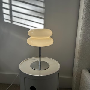 May include: A modern, white bedside table with a chrome lamp. The lamp has a stacked, rounded shade that emits a warm glow. The table is cylindrical with a single drawer and a circular top.