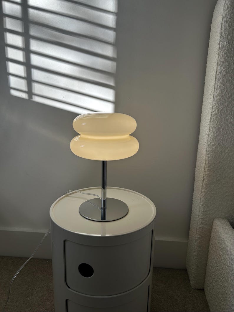 May include: A modern, white bedside table with a chrome lamp. The lamp has a stacked, rounded shade that emits a warm glow. The table is cylindrical with a single drawer and a circular top.