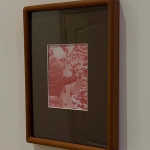 May include: A rectangular wooden picture frame with a dark brown mat and a red-toned photograph. The frame is mounted on a light gray wall. The photograph depicts a person and a child.