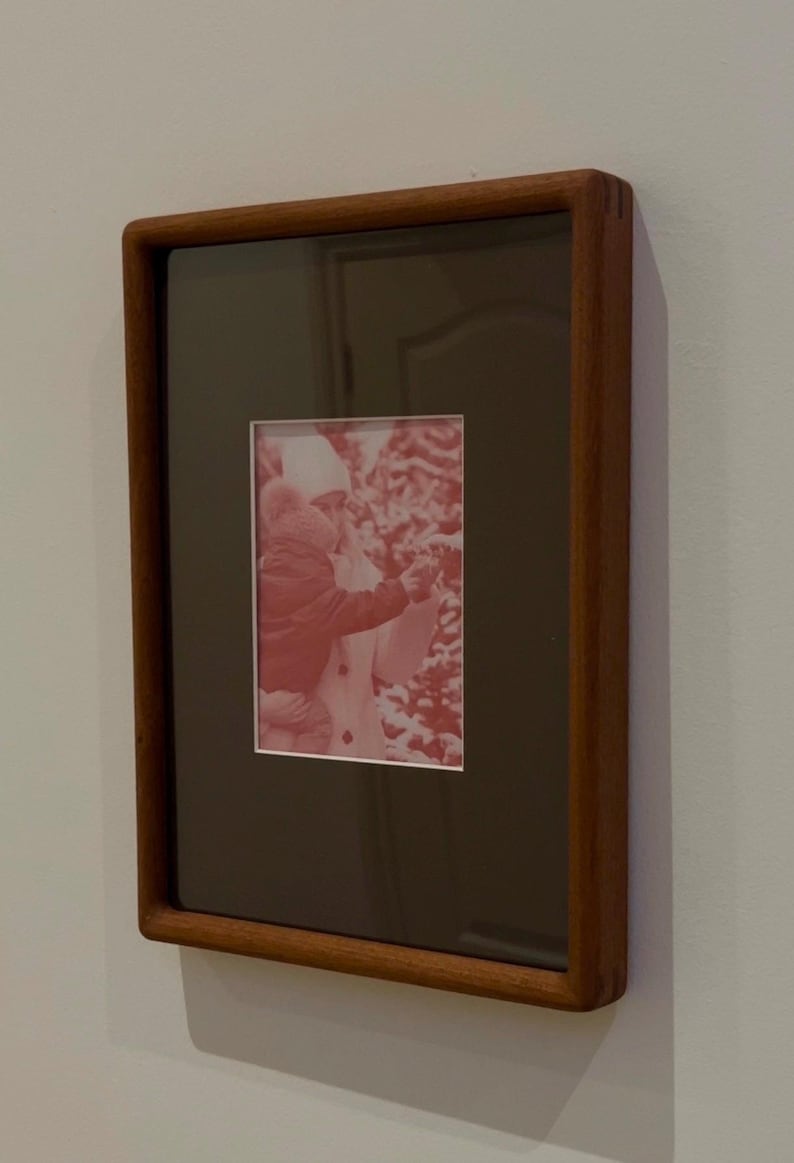 May include: A rectangular wooden picture frame with a dark brown mat and a red-toned photograph. The frame is mounted on a light gray wall. The photograph depicts a person and a child.