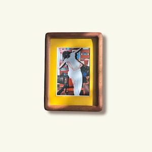 Puede incluir: Un marco de fotos rectangular de madera con acabado marrón oscuro y un paspartú amarillo brillante. El marco contiene una fotografía que muestra a una persona con ropa blanca de pie en una habitación con paredes rojas y varios objetos.