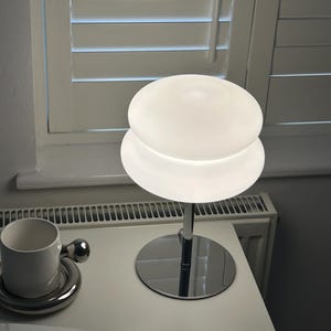 May include: A modern, white table lamp with a chrome base and stem. The lamp features a two-tiered, frosted glass shade. A white ceramic cup and saucer with a chrome handle are also on the table.