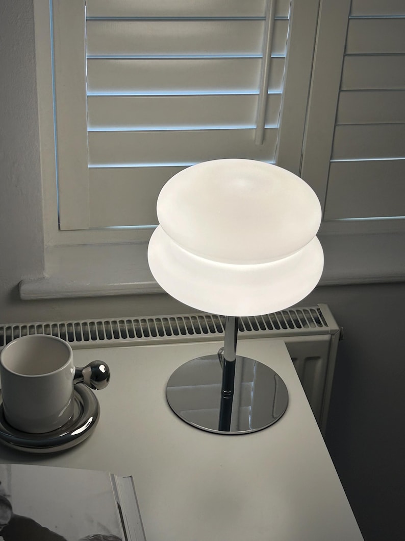 May include: A modern, white table lamp with a chrome base and stem. The lamp features a two-tiered, frosted glass shade. A white ceramic cup and saucer with a chrome handle are also on the table.