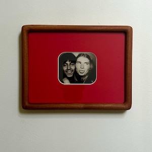 May include: A framed black and white photograph of two people making silly faces. The photo is set against a red background within a wooden frame. The frame is rectangular with rounded corners, and the photo is mounted on a white wall.