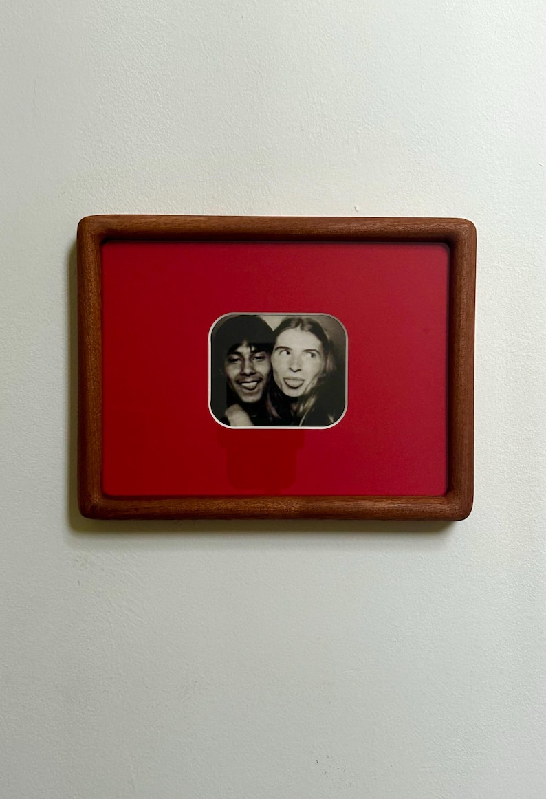 May include: A framed black and white photograph of two people making silly faces. The photo is set against a red background within a wooden frame. The frame is rectangular with rounded corners, and the photo is mounted on a white wall.
