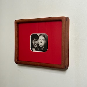 May include: A framed black and white photograph featuring two people with expressive faces. The photo is set against a vibrant red background within a square, wooden frame. The frame is mounted on a white wall.