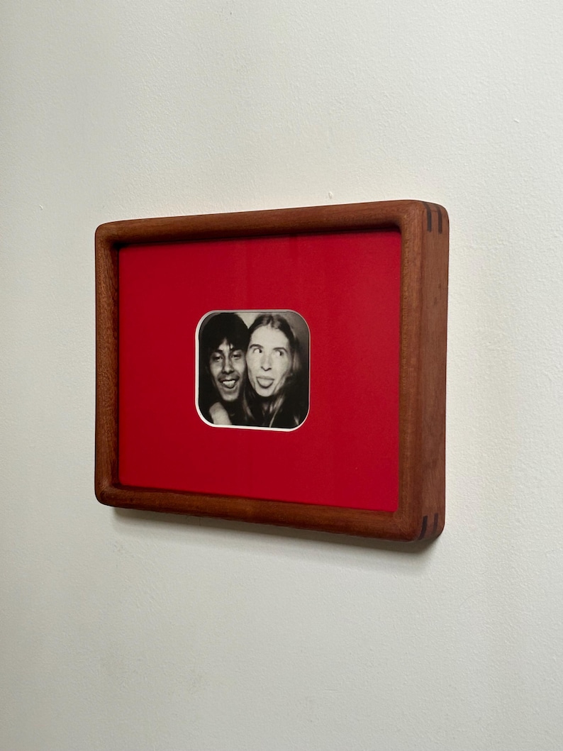 May include: A framed black and white photograph featuring two people with expressive faces. The photo is set against a vibrant red background within a square, wooden frame. The frame is mounted on a white wall.