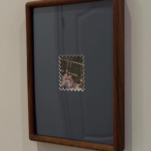 May include: A framed photograph with a dark wood frame. The photo is a small, postage stamp-like image of a family on a deck. The background of the frame is a muted blue-gray color, and the frame is mounted on a white wall.