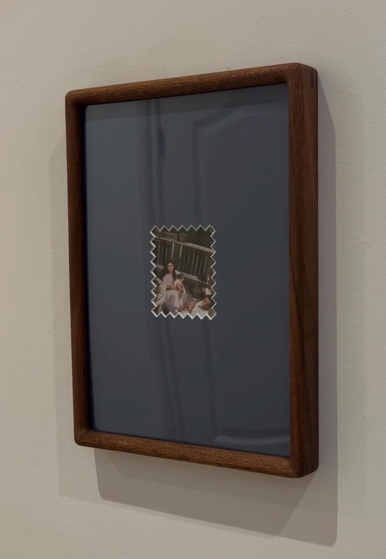 May include: A framed photograph with a dark wood frame. The photo is a small, postage stamp-like image of a family on a deck. The background of the frame is a muted blue-gray color, and the frame is mounted on a white wall.