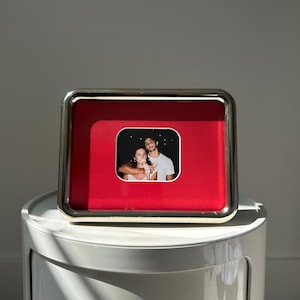 May include: A rectangular silver-toned picture frame with a vibrant red velvet-textured matting. Centered within the frame is a small, rounded square photograph depicting two people eating ice cream against a dark, starry night sky background.