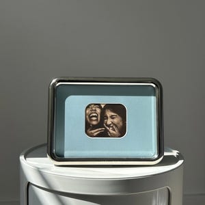 May include: A rectangular silver-toned picture frame with a light blue matting border. The frame contains a small, centered black and white photograph showing two individuals laughing with their hands near their faces in a candid expression.