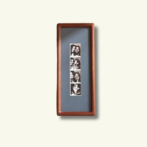 May include: A rectangular wooden frame with a dark brown finish containing a vertical strip of four black and white photo booth pictures. The photos are mounted on a solid gray mat board centered within the frame, displaying two people posing together.