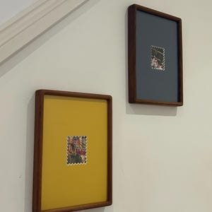 May include: Two framed artworks hang on a white wall. Each frame has a dark wood finish. One frame has a yellow background, the other is gray. Both frames feature a postage stamp in the center.