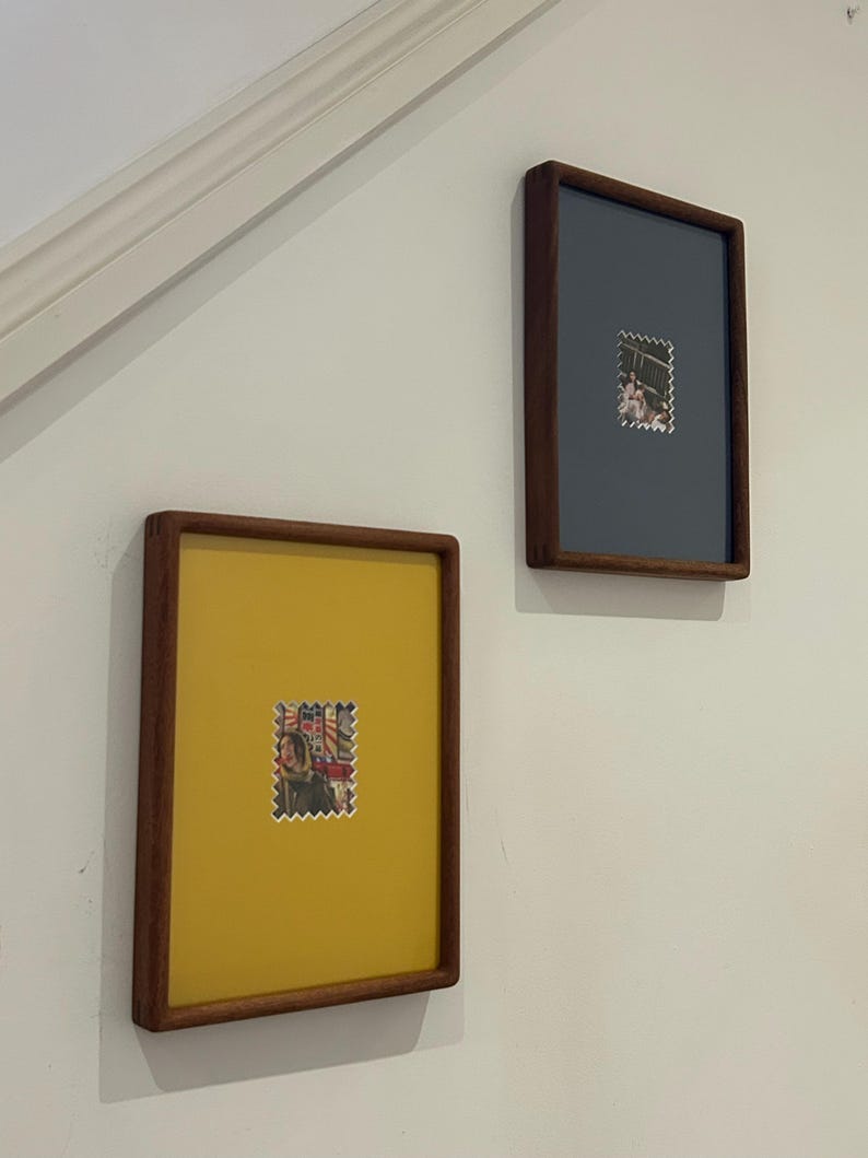 May include: Two framed artworks hang on a white wall. Each frame has a dark wood finish. One frame has a yellow background, the other is gray. Both frames feature a postage stamp in the center.