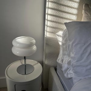 May include: A white, two-tiered table lamp with a chrome stem sits atop a white, rounded nightstand with a drawer. The lamp's shade has a stacked, disc-like design. A bed with a textured headboard is in the background.