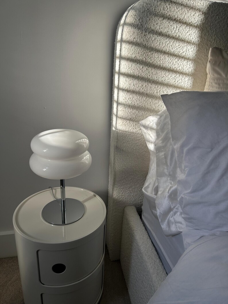 May include: A white, two-tiered table lamp with a chrome stem sits atop a white, rounded nightstand with a drawer. The lamp's shade has a stacked, disc-like design. A bed with a textured headboard is in the background.