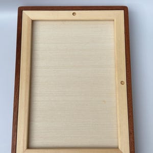 May include: A rectangular wooden frame with a light-colored interior and a dark brown border. The frame has a natural wood grain appearance and two small holes on the top and side.