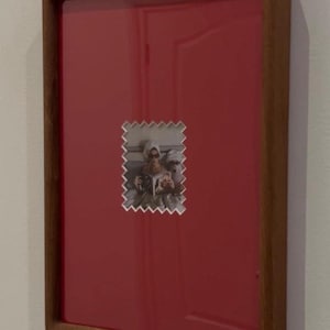 May include: A framed photograph with a red background and a wooden frame. The photograph is a small, postage stamp-shaped image of two people. The frame is rectangular and has a dark wood finish.