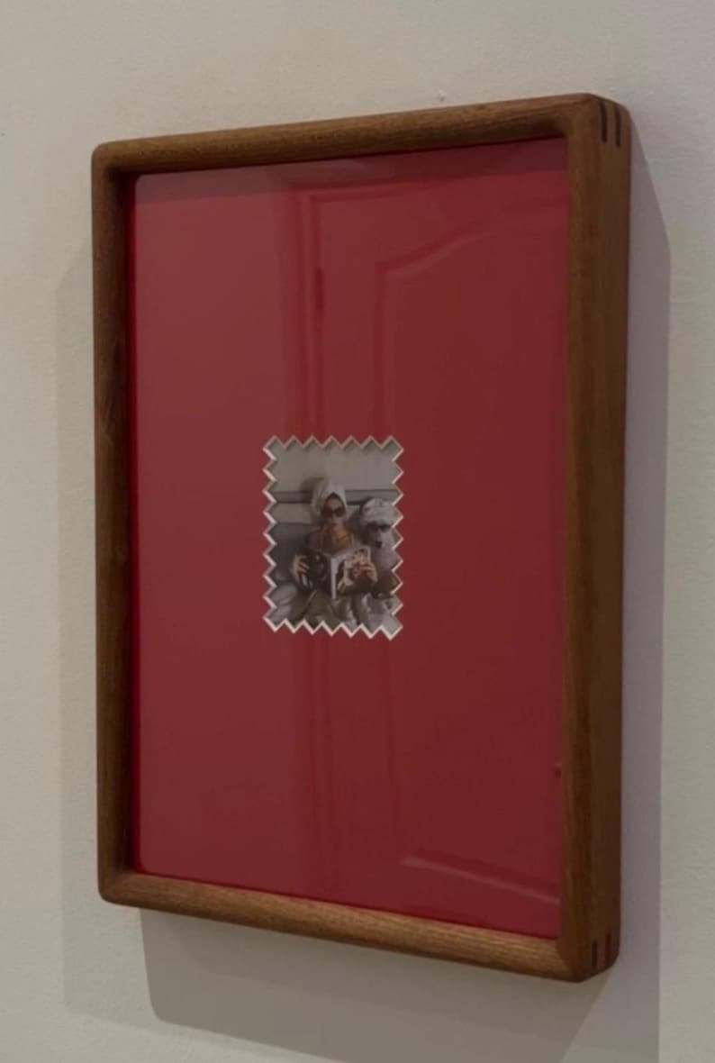 May include: A framed photograph with a red background and a wooden frame. The photograph is a small, postage stamp-shaped image of two people. The frame is rectangular and has a dark wood finish.